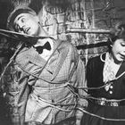 Zwei Personen in einer Schwarz-Weiß-Szene sind an eine Backsteinwand gefesselt. Beide tragen formelle Kleidung.