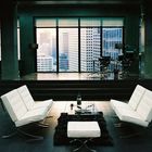 Ein moderner Raum mit weißen Sofas, Tisch, Teppich und großer Fensterfront mit Blick auf Wolkenkratzer im Hintergrund.