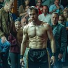 Ein Mann mit muskulösem Körper steht in einer Arena. Zuschauer um ihn herum, einige schauen interessiert zu.