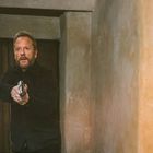 Ein Mann mit Bart und dunkler Kleidung hält eine Pistole in einem schmalen, beige-braunen Raum.