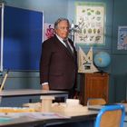 An einer Tafel steht "les enfants". Ein Mann im Anzug steht in einem Klassenzimmer mit Poster und Globus im Hintergrund.