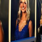 Eine Frau betrachtet große Porträts von lächelnden Frauen in eleganten Kleidern an einer Wand.