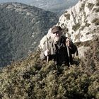 Ein Mann in traditioneller Kleidung mit Hut und Stöcken steht im dichten Gebüsch vor einer beeindruckenden Berglandschaft.
