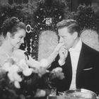 Eine elegant gekleidete Frau und ein Mann sitzen auf verzierten Stühlen, der Mann küsst der Frau die Hand.