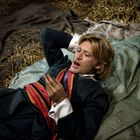 Ein Mann mit blonden Haaren liegt entspannt auf einem Bett aus Stroh und Stoff, trägt historische Kleidung.