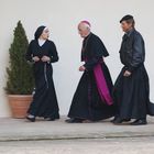 Keine sichtbaren Texte. Eine Nonne, ein Priester und ein Mann in Lederjacke gehen nebeneinander auf einem Innenhof.