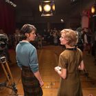 Zwei Frauen stehen auf einem Filmset mit Kameras und Scheinwerfern. Im Hintergrund sind Crew-Mitglieder und Requisiten.