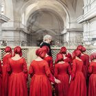 Frauen in roten Kleidern mit Hauben stehen in einer historischen, ornamentalen Kirche. Ein Mann mit weißer Perücke dabei.
