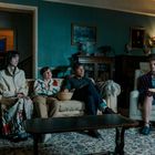 Es gibt keine sichtbaren Texte. Vier Personen sitzen auf Sofas in einem gemütlichen Wohnzimmer. Eine Person ist angekettet.