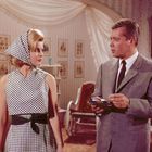 Ein Mann im Anzug und eine Frau in einem karierten Kleid stehen in einem elegant eingerichteten Raum.