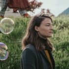 Eine Frau mit langen Haaren im Profil, vor grünem Gras. Seifenblasen schweben in der Luft. Hintergrund: Berge und Bäume.