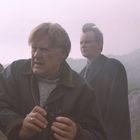 Zwei Männer im Freien; einer hält ein Fernglas, der andere steht im Hintergrund, beide blicken ernst.