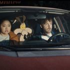 Zwei Menschen sitzen in einem Auto. Zwischen ihnen eine Comic-Figur mit großen, schockierten Augen.