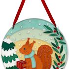Illustration: Ein Eichhörnchen mit Schal hält ein Geschenk vor einem Schneebaum, Schneefall, roten Beeren und grünem Blattwerk.