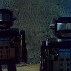 Zwei dunkle Retro-Roboter stehen vor einer strukturierten grauen Wand.