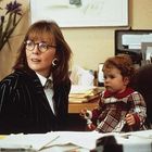Eine Frau mit Brille in einem Büro, trägt einen schwarzen Anzug. Ein Kind in einem karierten Kleid sitzt daneben. Büro mit Akten.