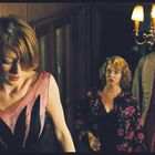 Keine Texte sind sichtbar. Drei Personen auf einer Treppe, beleuchtet von einer Deckenleuchte. Eine Frau vorne, zwei dahinter.