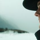 Ein Mann im Profil mit Hut und Mantel, blickt nach rechts in eine winterliche Landschaft mit Schnee und Bergen im Hintergrund.
