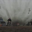 Mehrere Motorradfahrer fahren auf schlammigem Boden von einem großen Explosionsbereich weg.