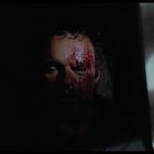 Kein Text vorhanden. Ein Mann steht im dunklen Raum, sein Gesicht ist blutverschmiert und teils im Schatten. Licht schimmert seitlich.