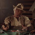 Ein älterer Mann mit Hut sitzt lässig am Tisch, hält einen Revolver. Auf dem Tisch liegen Pokerchips und Gläser.