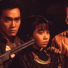 Ein Mann hält eine Waffe an den Kopf einer Frau. Ein weiterer Mann schaut besorgt drein. Dramatische Szene.