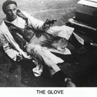 Text: "THE GLOVE". Mann in hellem Mantel liegt auf dem Boden, hält eine Pistole, schaut überrascht. Kartons im Hintergrund.