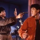 Zwei junge Männer, einer in brauner Jacke mit erhobener Pistole, der andere in orangefarbener Jacke, in einem Werkstattraum.