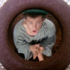 Es gibt keinen sichtbaren Text. Eine Person in einem grauen Gewand schaut durch ein kreisförmiges Objekt nach oben.