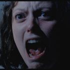 Eine Frau mit weit geöffnetem Mund und entsetzten Augen in einem düsteren Raum mit gepunkteter Wand im Hintergrund.