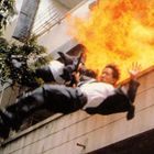 Zwei Männer in Anzügen stürzen von einem brennenden Gebäude, Flammen im Hintergrund, Chaos und Dramatik.