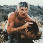 Zwei Männer im Freien. Der vordere Mann hält den anderen von hinten fest. Umgeben von Felsen und Gras.