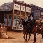 Schilder mit "Tommy Barber Shop" und "Bath". Zwei Männer auf Pferden in einer Westernstadt.
