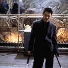 Person in dunkler Kleidung steht vor einer Wand mit Steinreliefs. Oben kauern Menschen in roten und blauen Anzügen.