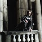 Eine Frau in schwarzer Lederkleidung kniet auf einem steinernen Geländer vor massiven Säulen, ihre Haltung wirkt gespannt.