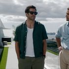Zwei Männer stehen auf einer Terrasse mit Meerblick. Einer trägt eine Sonnenbrille und ein grünes Hemd, der andere ein hellblaues Hemd.