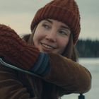 Eine lächelnde Person mit brauner Strickmütze und Handschuhen im Winter vor einem gefrorenen See im Hintergrund.