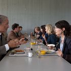 Zwei Personen sitzen sich in einer Kantine an einem Tisch gegenüber, umgeben von anderen Gästen.