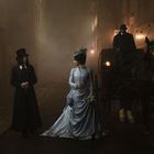 Im Hintergrund ein Schild mit schwer lesbarem Text. Personen in viktorianischer Kleidung neben einer Kutsche.