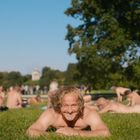 Ein Mann liegt lächelnd auf dem Bauch im grünen Gras, umgeben von weiteren Menschen und Bäumen im Hintergrund.