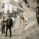 Vier Personen auf einer surrealen, verschachtelten Treppenkonstruktion mit Spiegelwänden.