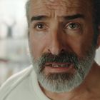 Ein Mann mit grauem Bart und besorgtem Blick steht in einem hellen Raum mit unscharfem Hintergrund.