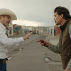 Zwei Männer stehen auf einer leeren Straße. Links trägt einer einen Cowboyhut und hebt die Hand, rechts zeigt der andere.