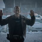 Ein Mann in Sheriff-Uniform hält die Hände hoch. Draußen Schnee, parkende Autos und blinkendes Polizeiauto.