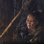 Eine Frau mit Kratzern im Gesicht kauert hinter einem Baum im Wald und schaut angespannt in die Ferne.