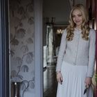 Eine Frau mit langen blonden Locken in einer grauen Strickjacke steht lächelnd in einer geöffneten Tür.