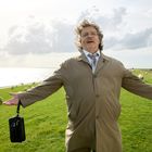 Ein Mann in einem beigen Mantel steht freudig auf einem Deich, Schafe grasen im Hintergrund, das Meer ist sichtbar.