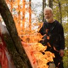 Ein älterer Mann steht im Wald, schaut auf ein großes Feuer, das von Holzstämmen und bunten Stoffen lodert.