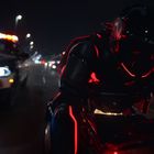 Ein futuristisches Motorrad mit einem Fahrer, umgeben von rotem Licht, wird von einem Polizeiauto mit Sirenen verfolgt.