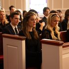 Eine Gruppe von Menschen sitzt dicht gedrängt in hölzernen Kirchenbänken, formell in dunkler Kleidung gekleidet.